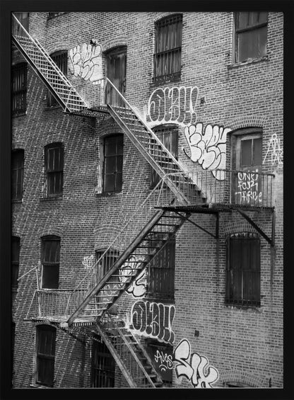 A black and white photograph of a brick apartment building with a prominent metal fire escape zigzagging across its facade. The walls are adorned with various styles of graffiti, and the windows are covered with bars, creating a gritty, urban scene. Decor