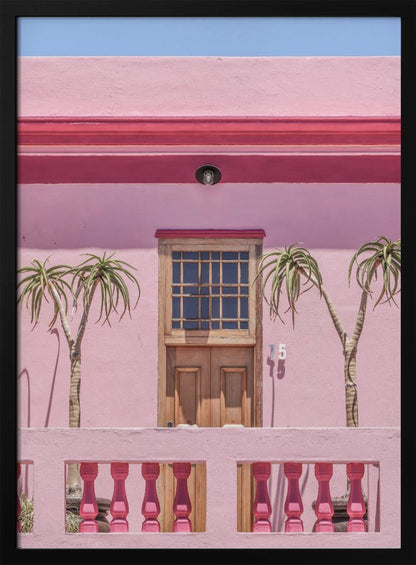 A brightly colored pink building stands under a clear blue sky, framed in black. A central wooden door with a paned window is flanked by two slender, palm-like trees. In the foreground, a pink balcony wall features fuchsia-colored balusters. Decor