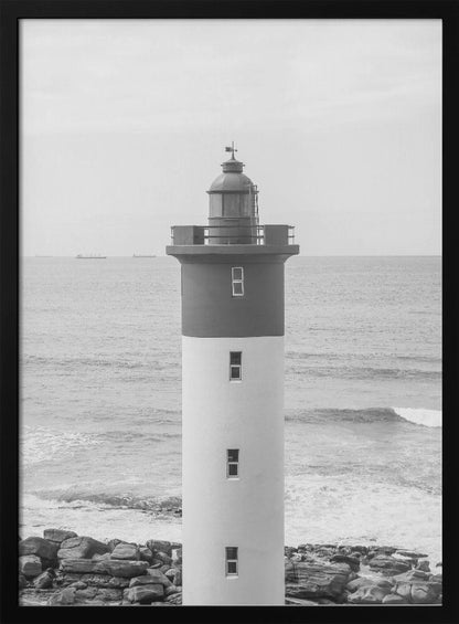 A framed, black and white photograph of a tall, cylindrical lighthouse standing on a rocky shore. The lighthouse is dark grey on its upper section and white on the lower, with the ocean and a grey sky in the background. Print