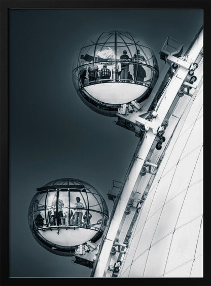 A dramatic low-angle, black and white photograph of two spherical glass pods of the London Eye Ferris wheel. Silhouettes of people are visible inside the pods against a dark, solid sky, with the massive white structure of the wheel on the right. Print