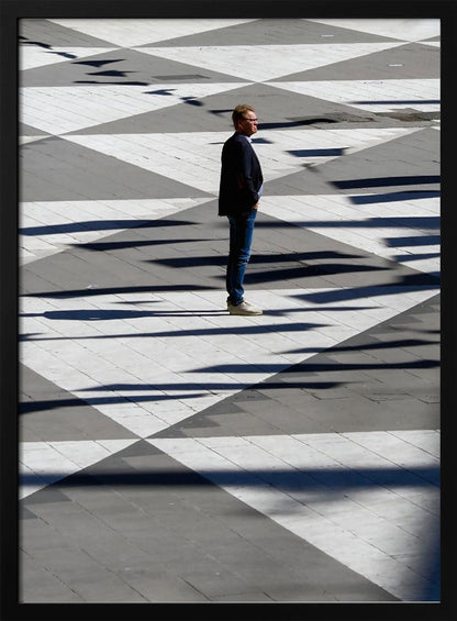A man in a dark jacket and blue jeans stands alone in profile on a geometrically patterned plaza. The ground is a striking pattern of grey and white triangles, crossed by long, dark shadows cast by the bright sun, creating a high-contrast, graphic composition. Wall Art
