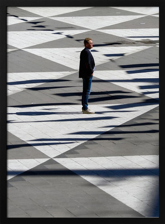 A man in a dark jacket and blue jeans stands alone in profile on a geometrically patterned plaza. The ground is a striking pattern of grey and white triangles, crossed by long, dark shadows cast by the bright sun, creating a high-contrast, graphic composition. Wall Art