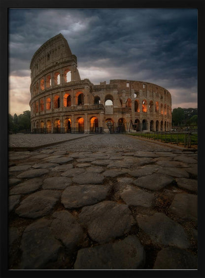 A low-angle view of the Roman Colosseum at dusk, with its arched windows glowing with warm orange light under a dramatic, stormy sky. An ancient cobblestone road leads towards the iconic amphitheater. Print
