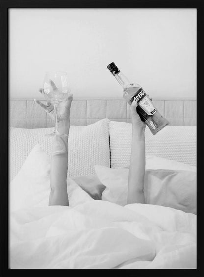 A black and white photograph of a person under white covers in bed, with only their arms visible. One hand holds up a bottle of Aperol, and the other holds up an empty wine glass. Decor