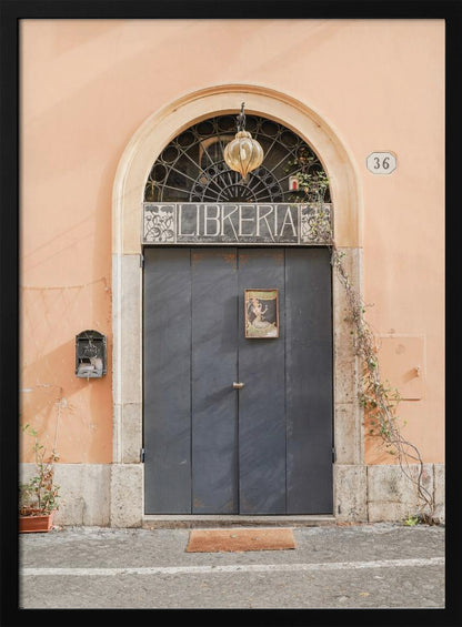 A charming European bookstore entrance featuring a large, dark grey arched door set against a peach-colored wall. Above the door, a sign reads 'LIBRERIA' below a decorative fanlight window and a hanging lantern. Decor