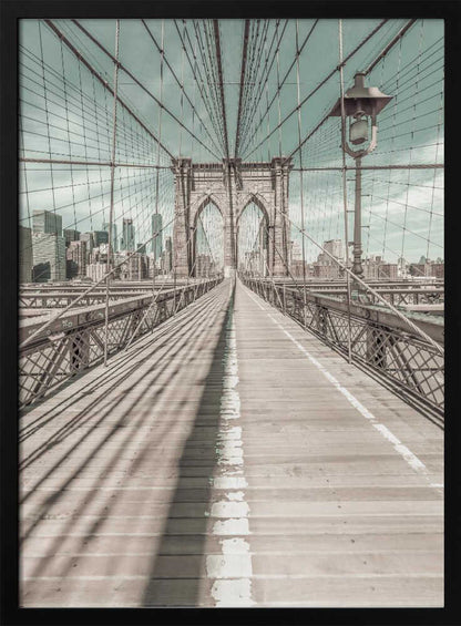 A vintage-style photograph looking down the center of the Brooklyn Bridge's wooden pedestrian walkway towards its iconic stone arches, with the New York City skyline visible in the background under a pale teal sky. The bridge's intricate network of suspension cables creates a strong sense of vanishing point perspective. Decor