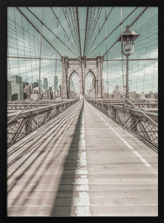 A vintage-style photograph looking down the center of the Brooklyn Bridge's wooden pedestrian walkway towards its iconic stone arches, with the New York City skyline visible in the background under a pale teal sky. The bridge's intricate network of suspension cables creates a strong sense of vanishing point perspective. Decor