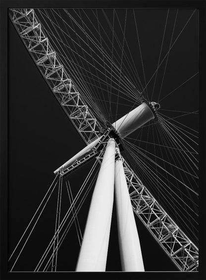 A dramatic low-angle, black and white photograph of a large Ferris wheel, focusing on the central hub and supporting structure. The image captures the white A-frame legs and the intricate lattice of the wheel's rim, with numerous tension cables radiating outwards against a solid black sky, creating a strong sense of perspective and geometric pattern. Artwork