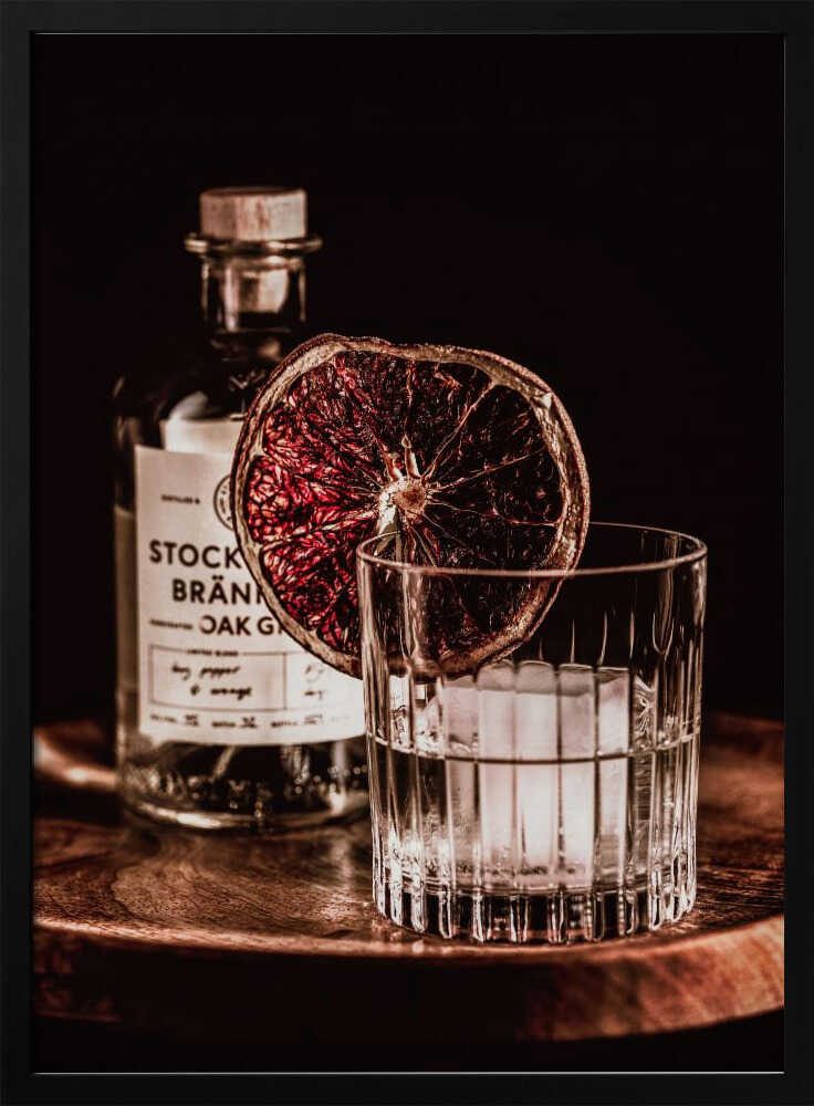 A moody still life photograph of a cocktail in a cut-crystal glass filled with ice. A dried blood orange slice garnishes the rim. In the soft-focus background, a bottle of gin rests on a dark wooden tray against a black backdrop. Wall Art