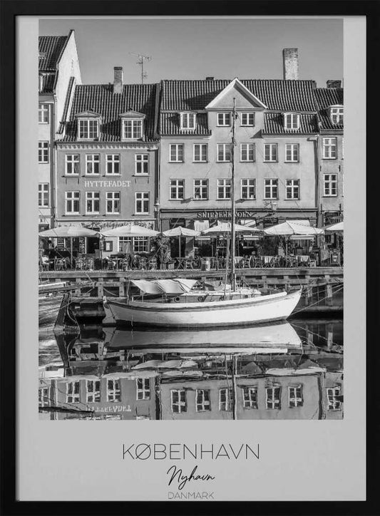 A black and white framed poster of Nyhavn in Copenhagen. The photograph features classic Danish buildings lining a canal, with a sailboat moored in the water. The buildings and boat are perfectly reflected in the still water. At the bottom, text reads 'KØBENHAVN', 'Nyhavn', and 'DANMARK'. Wall Art