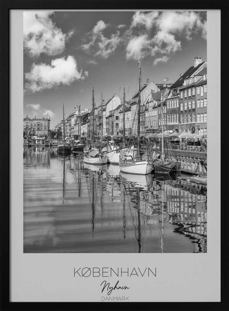A black and white poster of Nyhavn harbor in Copenhagen. The photograph shows traditional sailboats and ships moored along a canal, with a line of historic, multi-story buildings on the far bank. The sky has fluffy clouds, and the entire scene is reflected in the calm water. The poster has a white border with the text 'KØBENHAVN', 'Nyhavn', and 'DANMARK' at the bottom, and is shown within a black frame. Poster