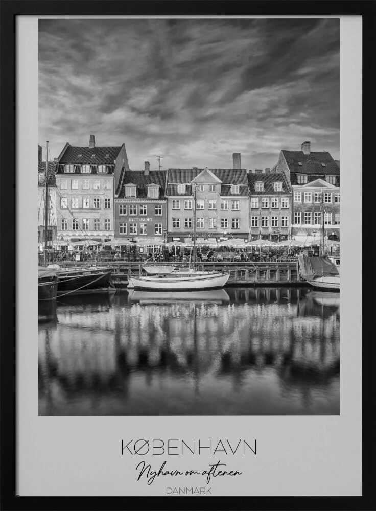 A framed black and white photograph of the Nyhavn harbor in Copenhagen, Denmark. The image shows a row of classic gabled houses along the waterfront, with several sailboats moored in the canal. The buildings and a dramatic cloudy sky are reflected in the water. At the bottom, a white border contains the text 'KØBENHAVN', 'Nyhavn om aftenen', and 'DANMARK'. Wall Art