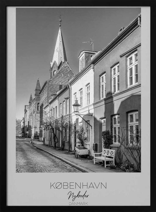 A black and white framed travel poster of a charming cobblestone street in Nyboder, Copenhagen. The photograph shows a sunlit row of historic houses with a prominent church steeple in the background. At the bottom of the poster, the words 'KØBENHAVN', 'Nyboder', and 'DANMARK' are written. Wall Art