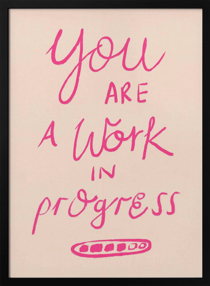 A framed art print featuring the motivational quote 'You are a work in progress'. The text is rendered in a vibrant pink, hand-lettered style against a soft beige background. The words 'You', 'a Work', and 'progress' are in a flowing script, while 'are' and 'in' are in a simple uppercase font. Wall Art