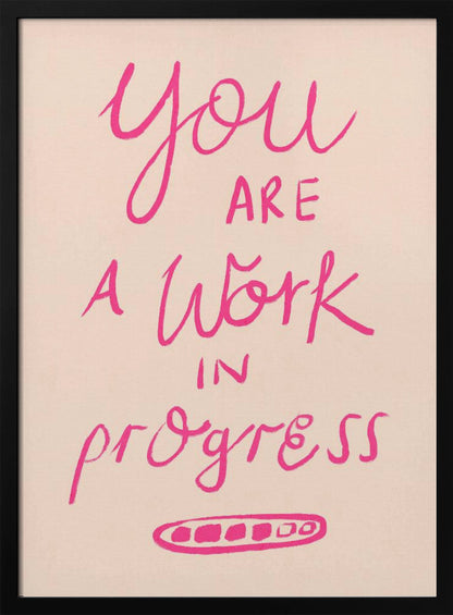 A framed art print featuring the motivational quote 'You are a work in progress'. The text is rendered in a vibrant pink, hand-lettered style against a soft beige background. The words 'You', 'a Work', and 'progress' are in a flowing script, while 'are' and 'in' are in a simple uppercase font. Wall Art