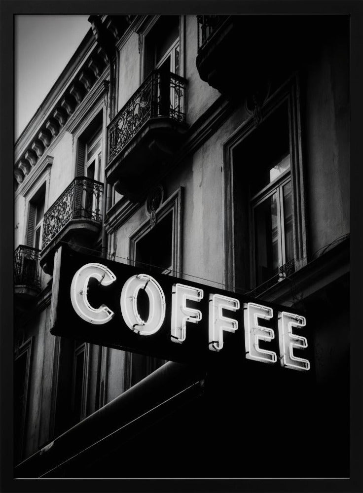 A dramatic, low-angle, black and white photograph of a bright white neon sign that reads 'COFFEE', mounted on a classic-style building with ornate balconies. Print
