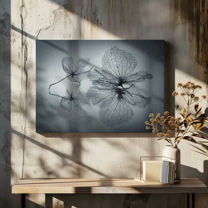 A framed, monochromatic photograph of two delicate skeleton flowers resting on a reflective surface. The intricate, web-like veins of the dried flowers are shown in close-up detail, and their faint reflections are visible below. The background is a soft, out-of-focus gray gradient. Artwork
