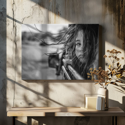 A close-up, black and white photograph of a young girl looking out of a moving vehicle. Her long hair is blowing wildly in the wind, partially obscuring her face as she peers intently at the camera with one eye. Her hand grips the edge of the open window. The background is blurred with motion, suggesting a journey. The photo is displayed in a silver frame. Poster