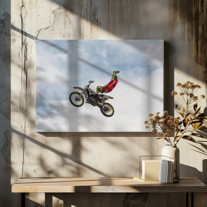 A motocross rider in red and yellow gear performs a high-flying stunt, arching their back while upside down in the air next to their dirt bike, set against a backdrop of a blue sky with white clouds and enclosed in a silver frame. Poster