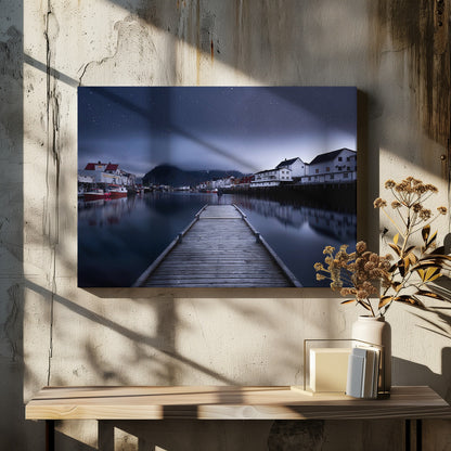 A long exposure photograph of a serene coastal town at night, viewed from a wooden pier that leads into calm, dark water. A lone person stands at the end of the pier, gazing at the town's reflected lights. Behind the village, a mountain looms under a breathtaking, star-filled sky. The image is presented within a silver-colored frame. Artwork