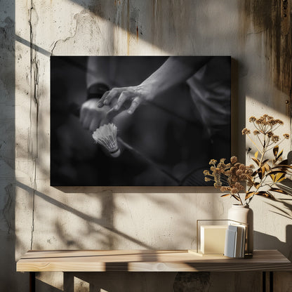 A dramatic black and white close-up photograph of a badminton player preparing to serve. The focus is on the player's hands, with one hand delicately positioned above a shuttlecock which is balanced on the strings of a racquet. Artwork