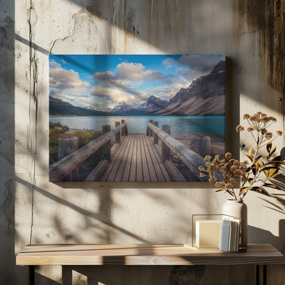 A wooden pier leads out onto a vibrant turquoise glacial lake, surrounded by towering, snow-dusted mountains under a dramatic cloudy sky. The image is presented in a silver frame. Artwork