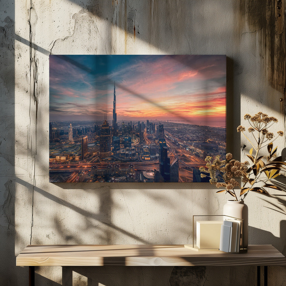 An aerial view of the Dubai skyline at sunset, with the Burj Khalifa prominently featured against a dramatic sky filled with orange, pink, and blue clouds. The sprawling city below is illuminated with golden lights from buildings and busy highways. Decor