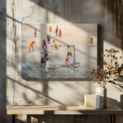 An overhead view of a group of boys and young men playing a spirited game of soccer on a large, dusty dirt field. A player in a bright orange uniform prepares to kick the ball towards a makeshift goal, defended by a goalie in a dark blue jersey. Other players in various colored shirts are scattered across the field, while a few young spectators watch from the sidelines. The entire image is enclosed in a silver frame. Wall Art