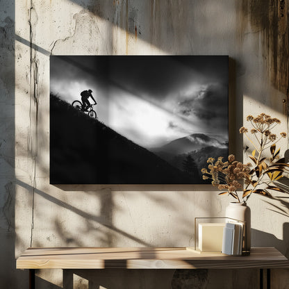 A striking black and white photo of a mountain biker's silhouette on a steep decline, set against a dramatic, light-filled cloudy sky with distant, misty mountains. Artwork