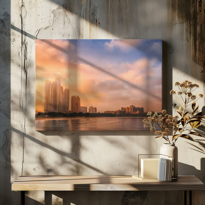 A framed photograph of the Abu Dhabi city skyline at sunset, featuring modern skyscrapers and the Emirates Palace hotel glowing in warm, orange light. The buildings are reflected in the calm water of the foreground, under a sky with soft orange and blue clouds. Poster