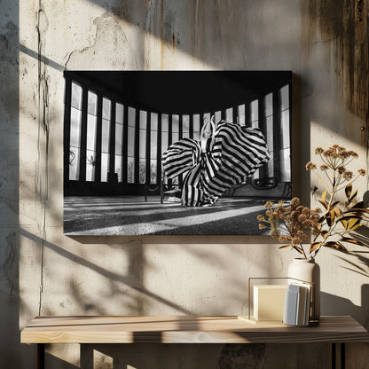 A dramatic black and white photograph of a woman in a voluminous, horizontally striped dress, captured mid-motion with the fabric swirling around her. She stands in a sunlit, circular room with tall, slatted windows that cast long shadows across the floor. Her arms are raised gracefully above her head, creating a striking and artistic pose. Artwork