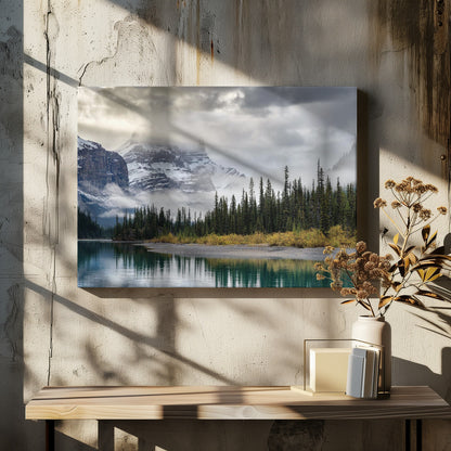 A tranquil mountain landscape featuring a still, teal lake reflecting a dense evergreen forest along its shore. In the background, majestic snow-covered peaks are shrouded in mist and dramatic gray clouds. Wall Art