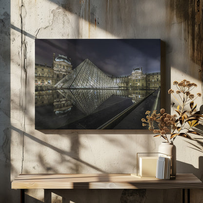 A dramatic night photograph of the illuminated Louvre Pyramid in Paris, its glass structure glowing against a dark, cloudy sky. The historic Louvre Palace is visible in the background, and the entire scene is perfectly reflected in the still water of a foreground pool. The image is presented within a silver frame. Artwork