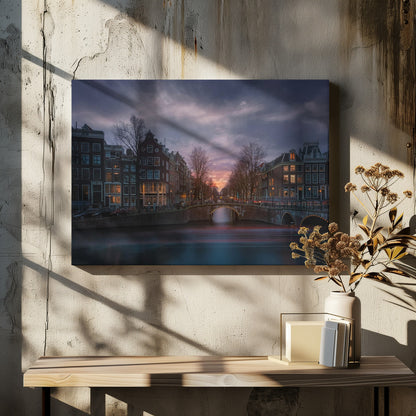 A dramatic long-exposure photograph of an Amsterdam canal at twilight. An arched bridge spans the water, lined by historic Dutch buildings with glowing windows. The sky is filled with dark, moody clouds above a fading orange and purple sunset, and red light trails from boats are blurred on the water's surface. Poster