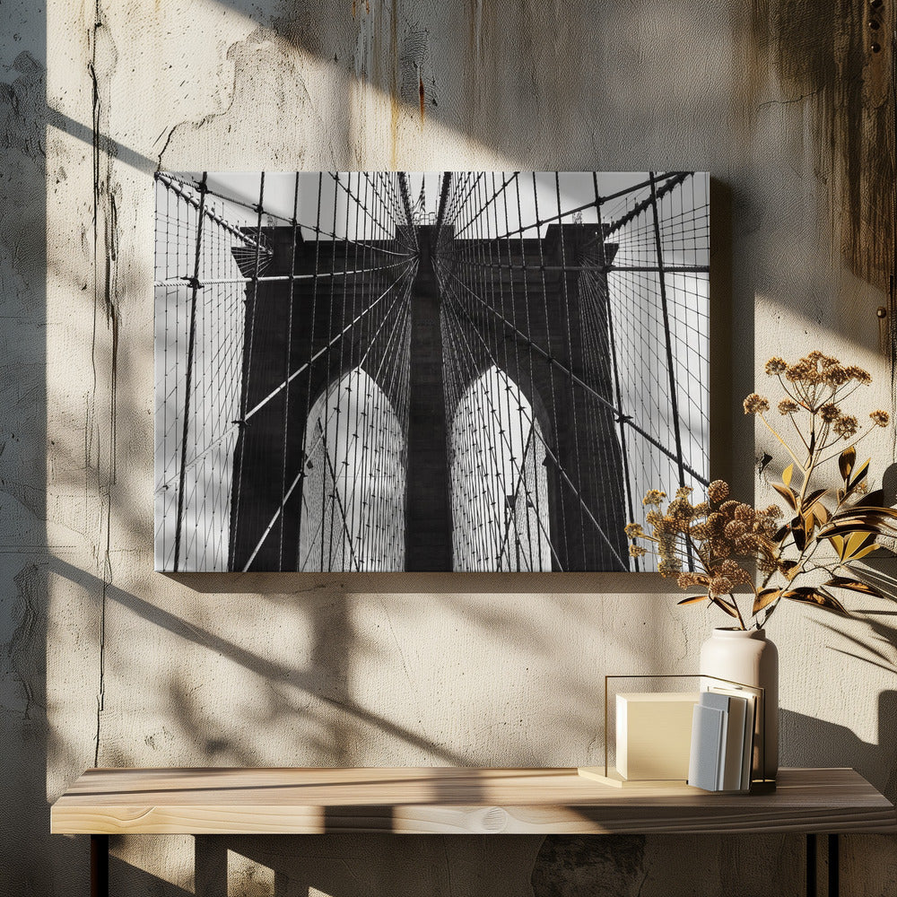 A low-angle, black and white photograph looking up at the iconic stone towers of the Brooklyn Bridge. The intricate web of suspension cables and vertical suspender ropes creates a symmetrical, geometric pattern against the light sky. The photo is enclosed in a silver frame. Print