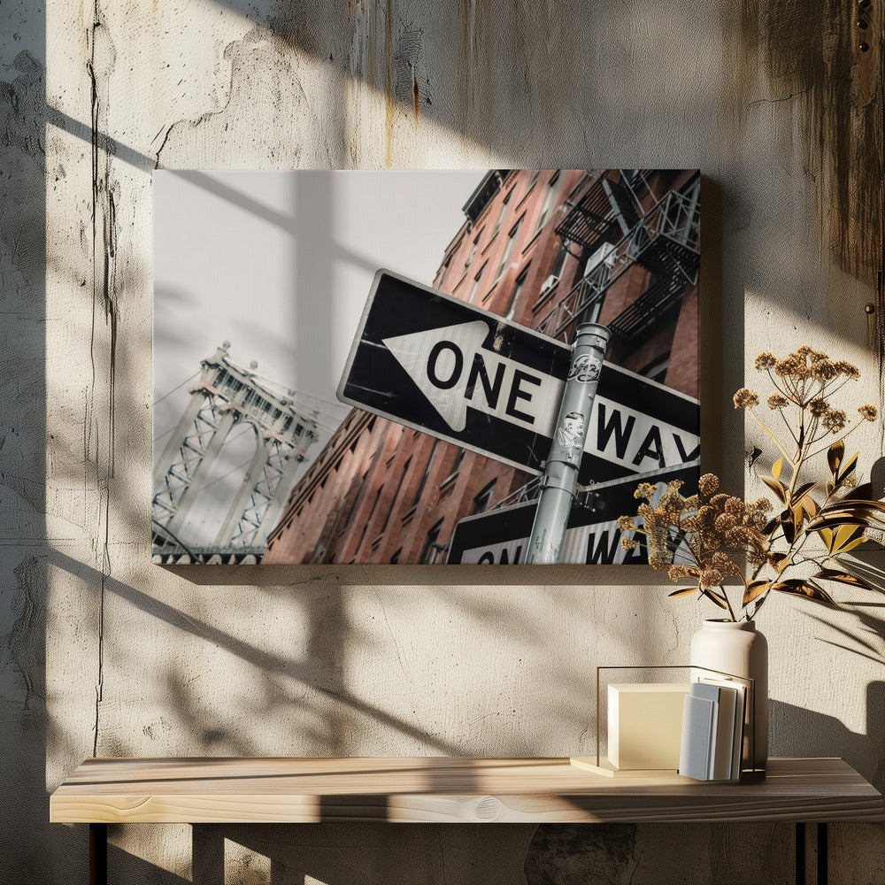 A low-angle shot of a black and white 'ONE WAY' street sign in the foreground, with a red brick building and the Manhattan Bridge visible in the background under an overcast sky, all enclosed in a silver frame. Artwork