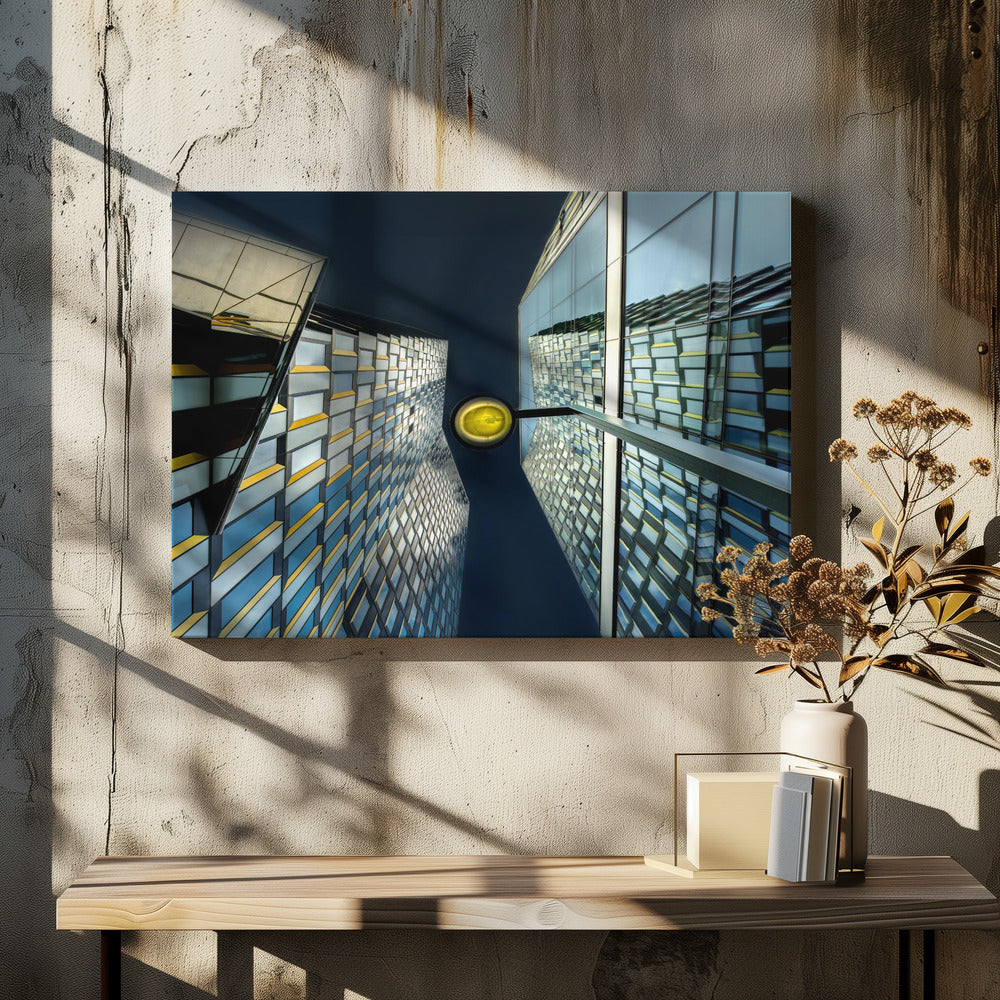 A worm's-eye view looking up between two modern glass skyscrapers with geometric blue, yellow, and silver panels, creating a symmetrical reflection against a dark blue sky, with a bright yellow streetlamp centered in the gap. Decor