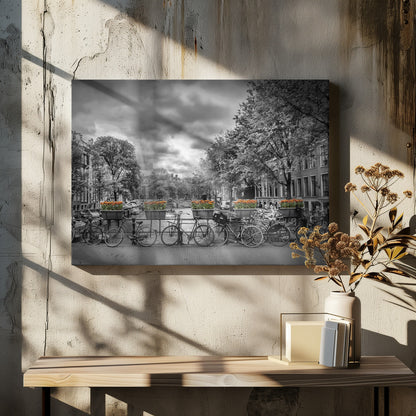 A selective color, black and white photograph of a classic Amsterdam canal scene, enclosed in a silver frame. The foreground shows a bridge lined with numerous bicycles and planters filled with vibrant orange tulips, which are the only colored elements. The background features canal houses, trees, and another bridge under a dramatic, cloudy sky with sunbeams. Print