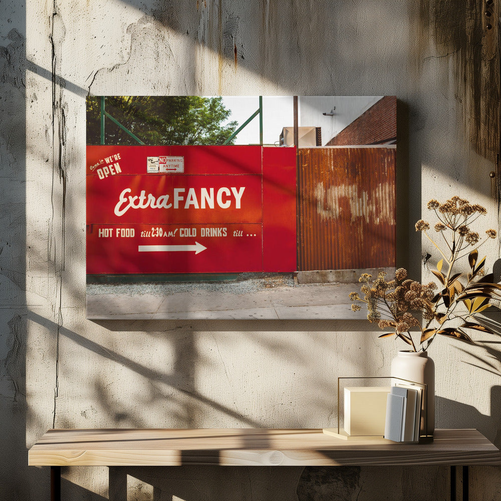 A framed photograph of a bright red outdoor sign for a restaurant named 'Extra Fancy'. The sign, painted with white retro-style text, advertises hot food and cold drinks and is situated next to a rusty, corrugated metal fence on a city sidewalk. Artwork