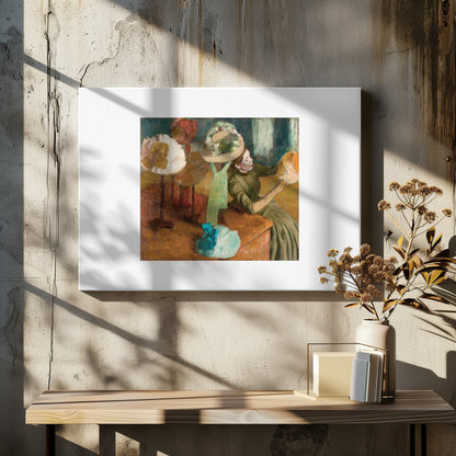 A framed print of Edgar Degas's Impressionist painting 'The Millinery Shop,' showing a woman in a green dress examining a hat in a shop filled with various fashionable hats on display stands. Print