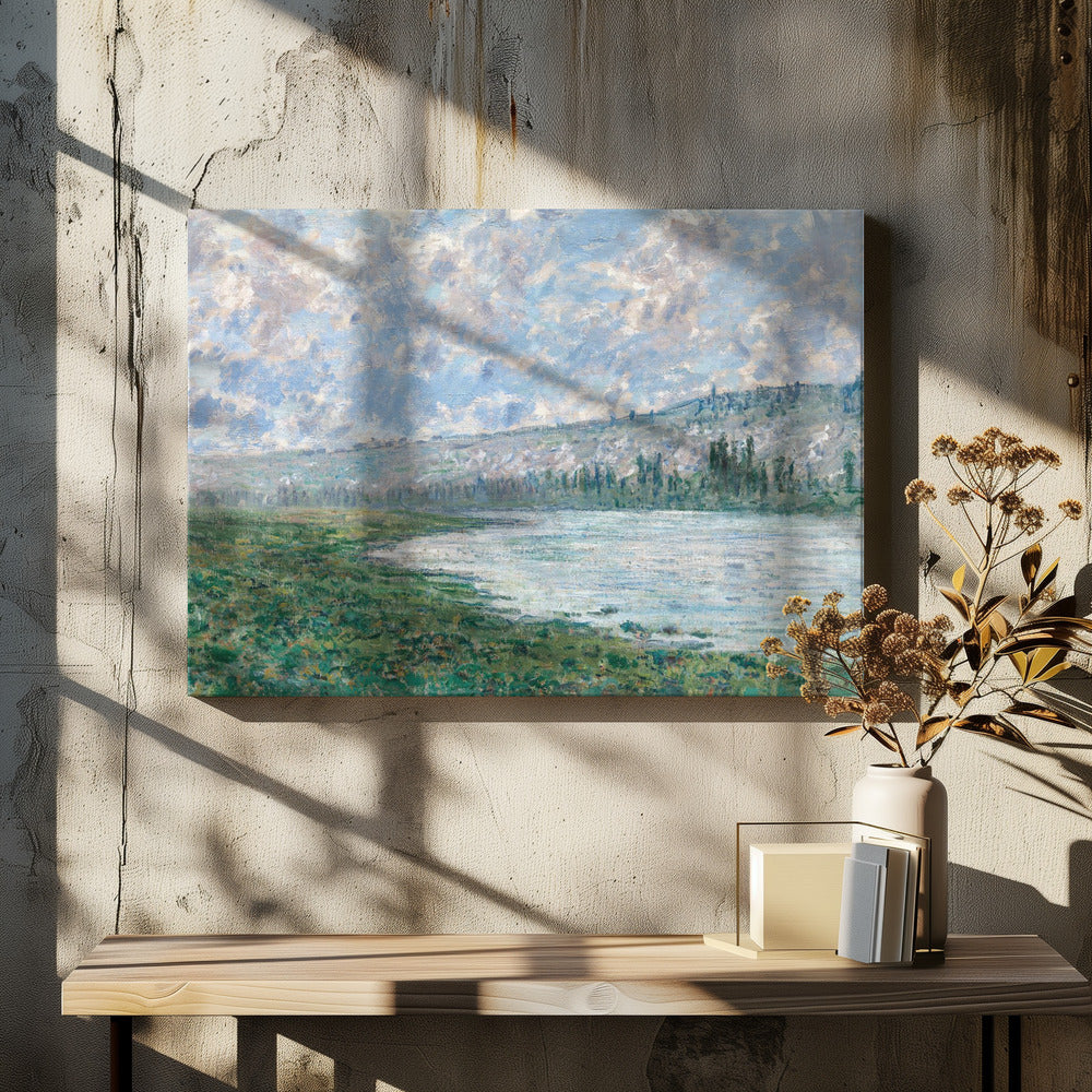 An Impressionist painting by Claude Monet, 'The Seine at Vétheuil', depicting a tranquil landscape. In the foreground, a lush green riverbank meets the calm, reflective water. A distant, soft-focus hillside rises under a vast sky filled with puffy white and grey clouds against a light blue background. The artwork is characterized by visible, textured brushstrokes and is displayed in a modern silver frame. Artwork