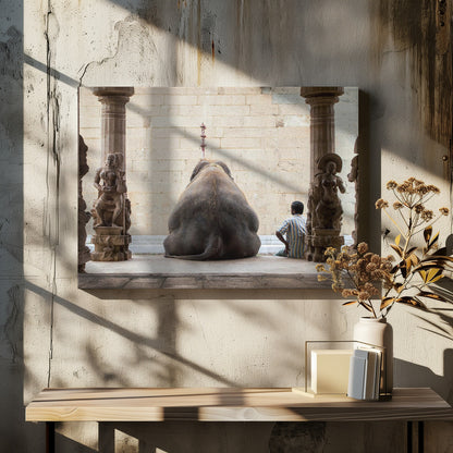 A framed photograph from behind a large elephant sitting on a stone floor inside a temple, flanked by ornate, carved stone pillars. A man in a striped shirt sits on the floor to the right of the elephant, looking towards it. Decor