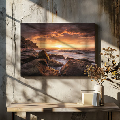 A framed photograph of a dramatic ocean sunset, with fiery orange clouds filling the sky. Large, dark rocks sit on the shore as long-exposure waves create a misty effect around them. Print