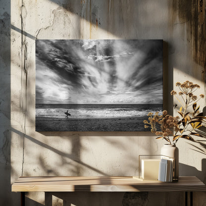 A dramatic black and white photograph of a lone surfer silhouetted against the sea, walking along a sandy beach. The sky is filled with spectacular, radiating clouds that stretch across the horizon, creating a powerful and moody atmosphere. The image is enclosed in a silver frame. Poster