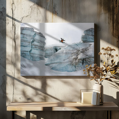 A framed photo of a skier in mid-air, jumping over a snowy crevasse between large, layered glacier formations under an overcast sky. The skier wears a black top and orange pants. Wall Art