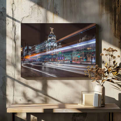 A dynamic long-exposure night photograph of the Metropolis Building in Madrid, with vibrant streaks of white, orange, and blue light from traffic swirling through the city streets. Artwork