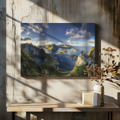 A breathtaking panoramic photograph of the Lofoten Islands in Norway from a high vantage point. A person stands on a rocky cliff with arms outstretched, overlooking a stunning landscape of deep blue fjords, lush green mountains, and the small fishing village of Reine below. The sky is a vibrant blue with scattered white clouds. Artwork