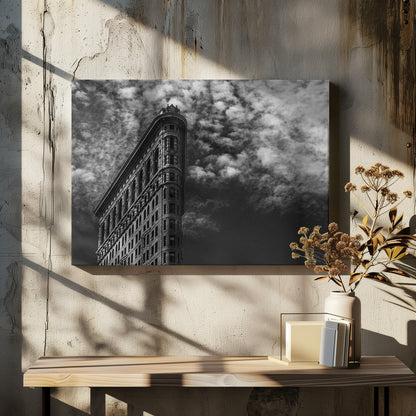 A dramatic low-angle black and white photograph of the Flatiron Building in New York City, with its sharp, triangular corner pointing towards a sky filled with fluffy, white clouds. The image is framed with a brushed silver border. Print