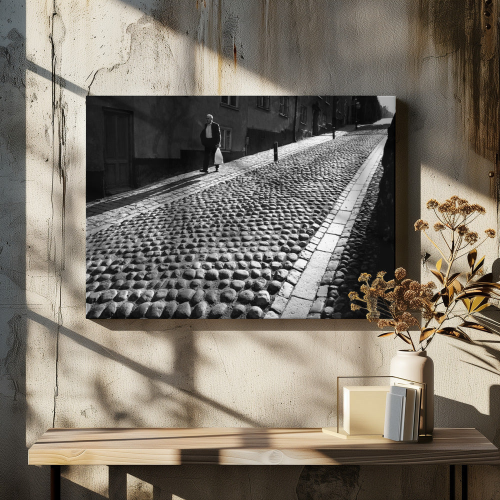 A framed, black and white photograph capturing an elderly man in a suit walking up a steep cobblestone street. The man carries a white bag and casts a long shadow behind him in the harsh sunlight. The street is lined with old, multi-story buildings, creating a classic European city scene. Poster
