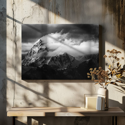 A dramatic black and white photograph of a snow-covered mountain peak being enveloped by thick, swirling clouds under a dark, stormy sky. The rugged slopes and sharp ridges of the mountain are highlighted by the snow, while a dark forest sits at the base. The entire image is presented within a textured silver frame. Print